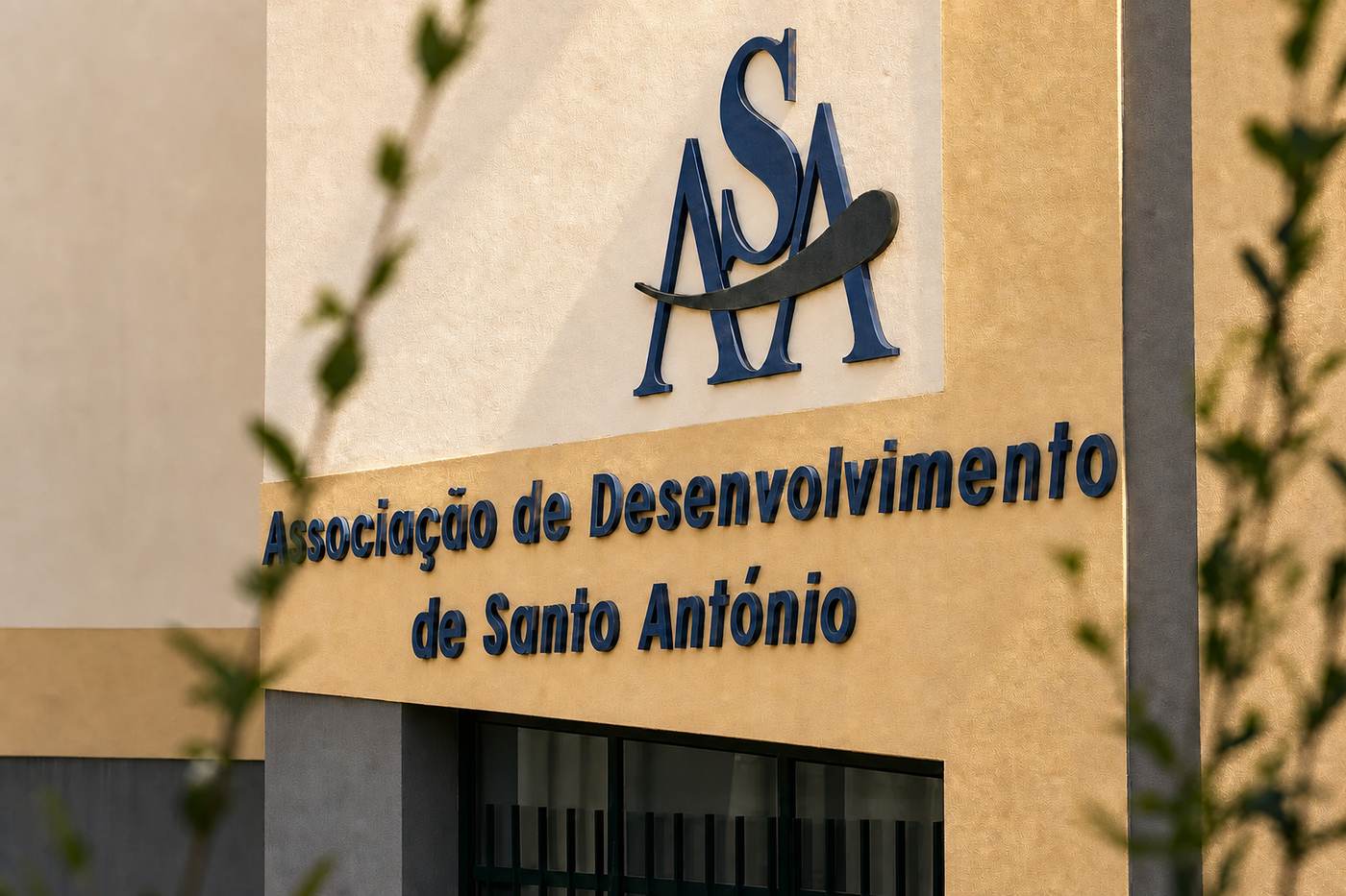 Fachada da sede da ASA ao fim da tarde. O logotipo da associação, em azul-escuro, está montado sobre um painel creme; por baixo, sobre a parede cor de mostarda, lê-se em letras azuis «Associação de Desenvolvimento de Santo António».