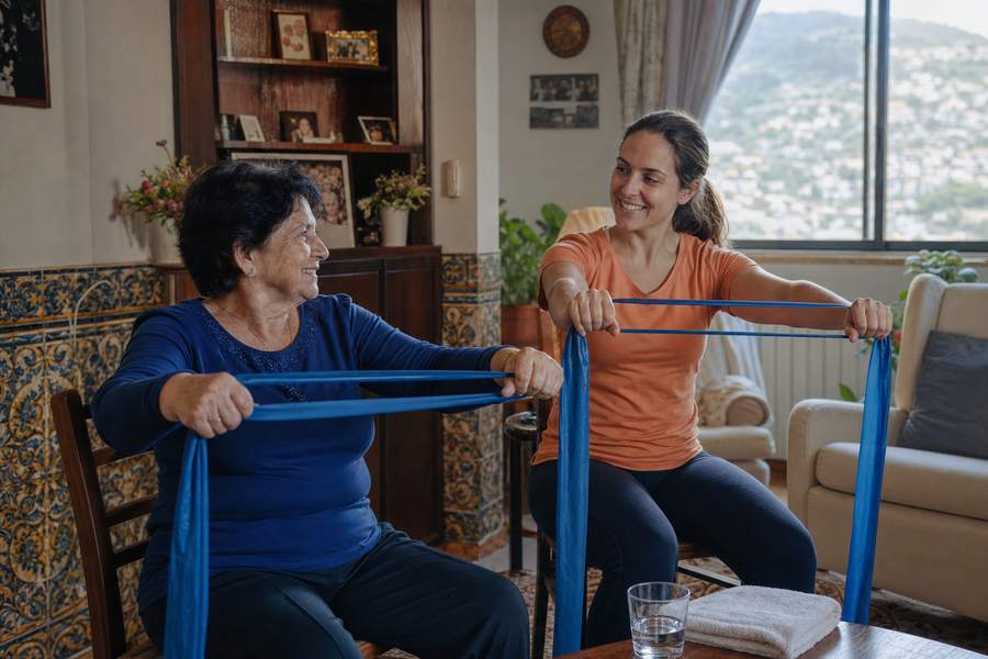 Uma senhora idosa e uma técnica da ASA a fazer exercício com faixa elástica, sentadas na sala de casa.